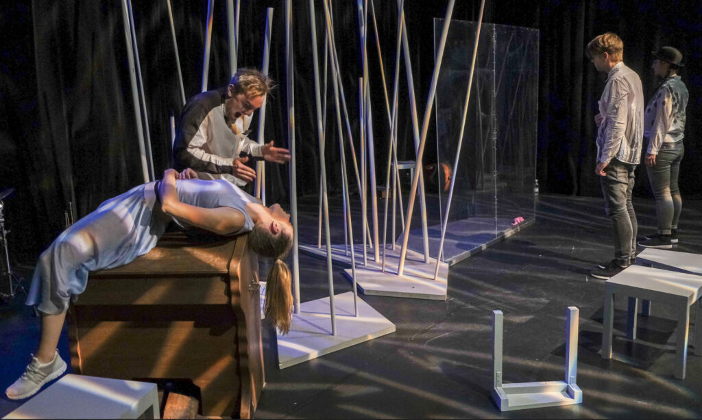 In einem Bühnenbild voller dünner Stangen lehnt sich ein Mann bedrohlich über den leblosen Körper einer Frau