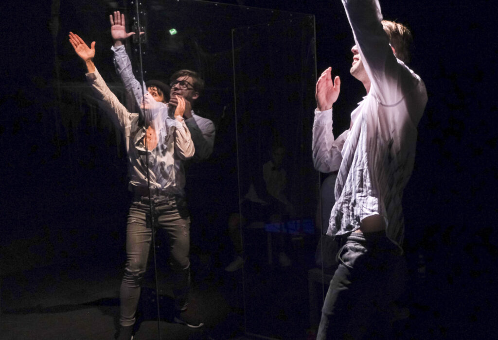 Ein Mann spiegelt sich in einer Glasscheibe während hinter der Glasscheibe eine Frau seine Pose imitiert