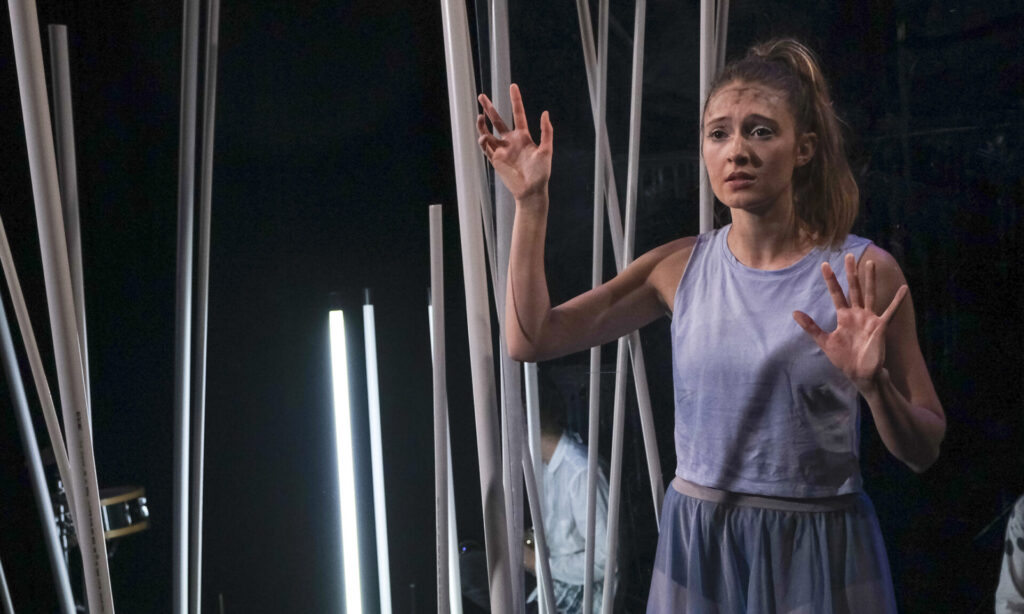 Eine Frau blickt sehnsuchtsvoll in die Ferne, hinter ihr ein Bühnenbild aus Stangen und Leuchtstäben.