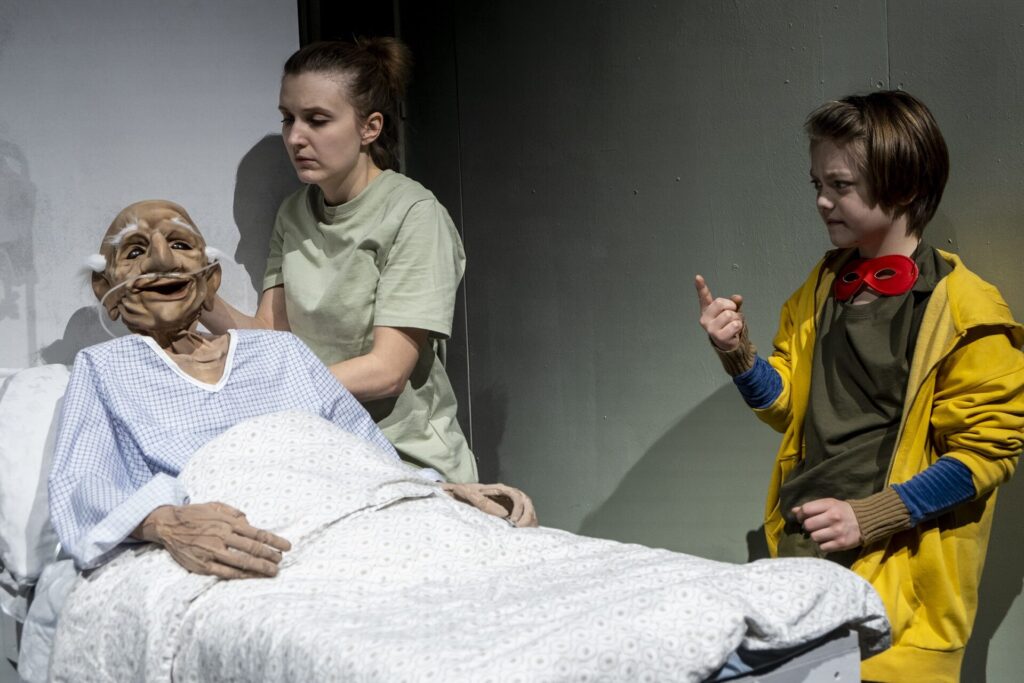 © Foto: Rainer Berson Junge steht an einem Bett, darin liegt die Puppe eines alten Mannes.