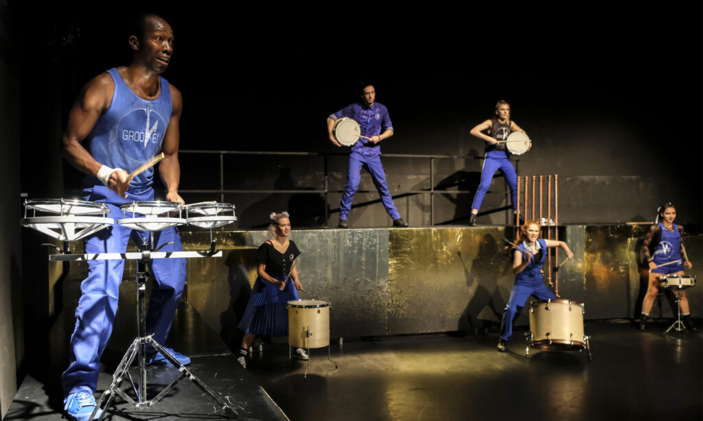 © Foto: Rainer Berson Sechs Performer:innen mit Trommeln stehen verteilt auf einer gold glänzenden Bühne