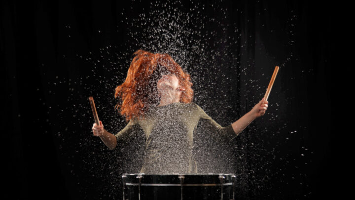 Eine rothaarige Frau schlägt dynamisch auf eine Trommel von der Wassertropfen wegspritzen